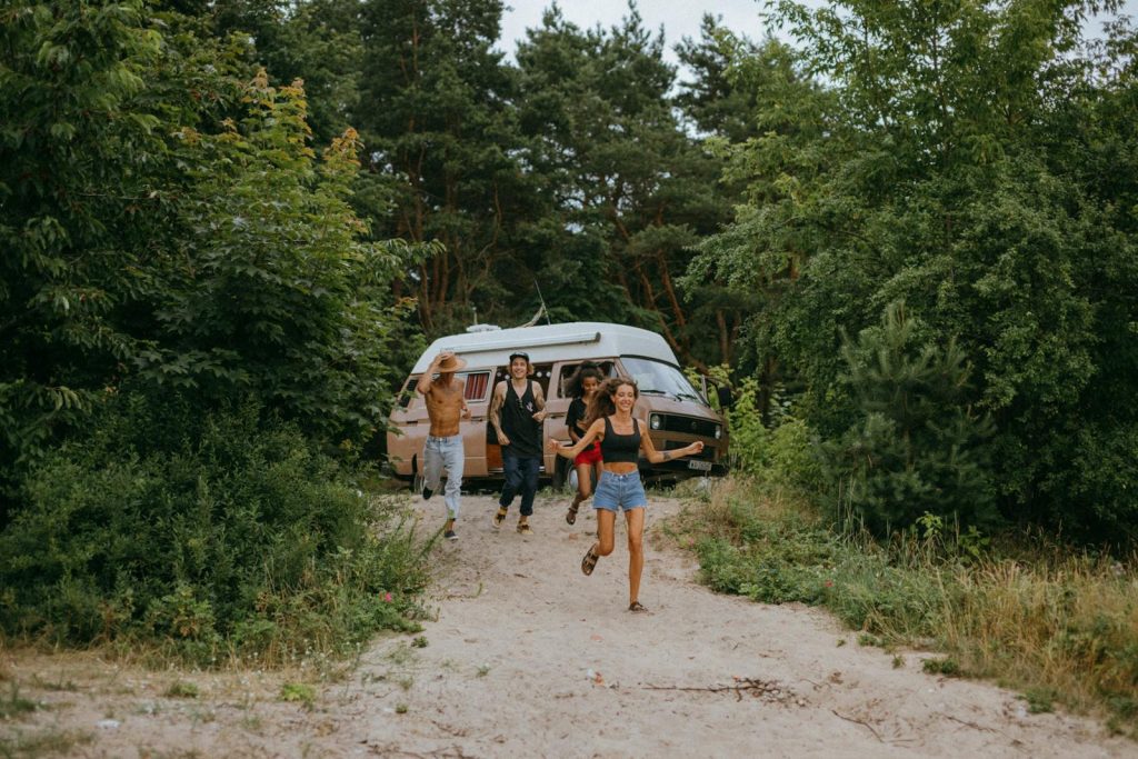 Group of friends running joyfully from a camper van in a lush forest setting during summer.